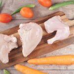 Overhead view of raw chicken, tomatoes, carrots, and okra on a chopping board.